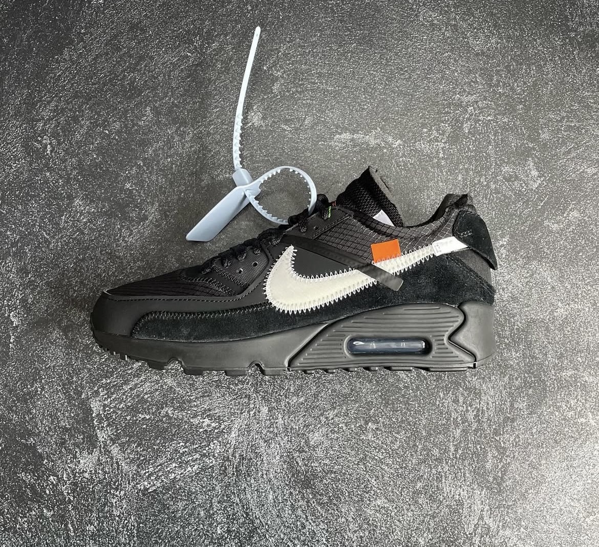 Black Nike Air Max Off-White sneaker with exposed stitching and signature zip tie detail on textured black and grey background.